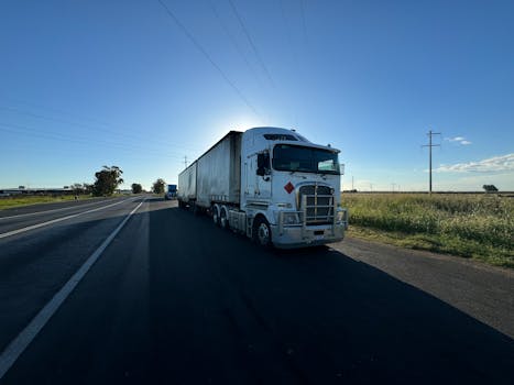 A silver semi truck driving on an open highway under a clear blue sky.